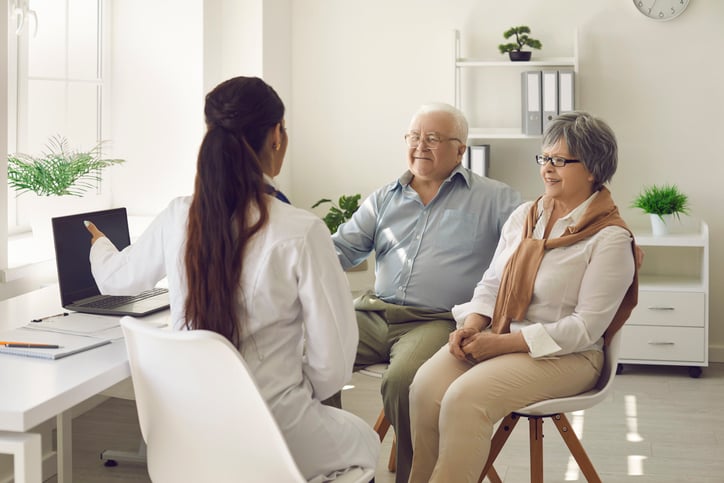 Happy mature couple and their doctor communicating while using laptop during medical appointment. Female doctor explains to senior patients test results pointing at laptop screen. Healthcare concept.